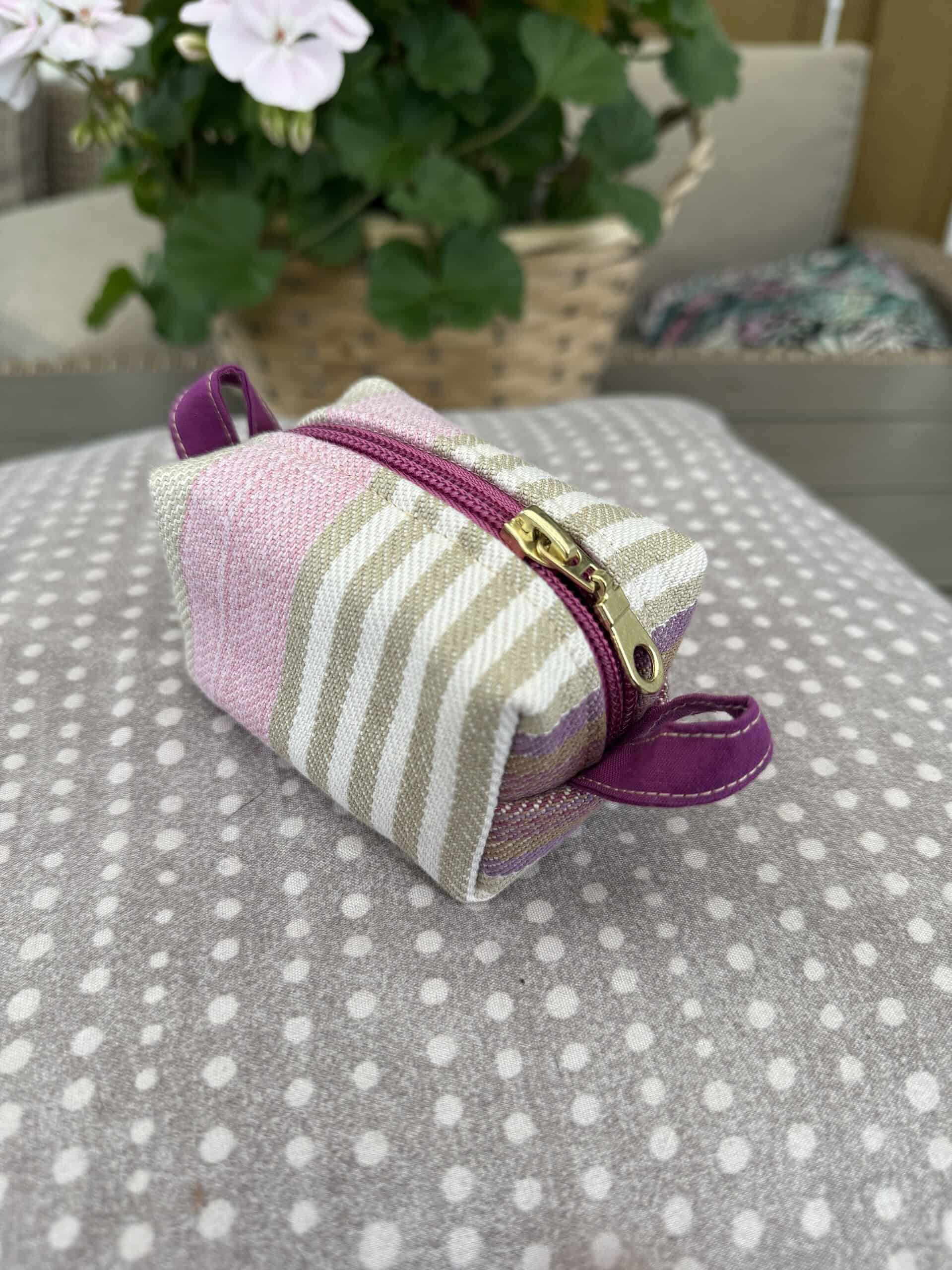 Mini tygpåse i pastellfärger med rosa, beige och lila ränder, guldfärgad dragkedja och handtag, perfekt för småsaker eller smycken.