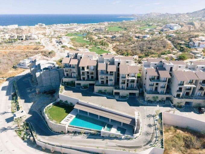 Aerial View Of A Luxury Golf Community With Ocean Views, Featuring Modern Residential Buildings, A Swimming Pool, And Scenic Landscape In The Background.