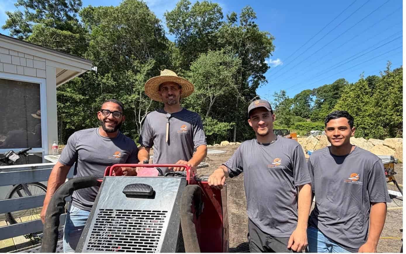 Coast Carpentry Home Group masonry crew in branded Coast Masonry shirts standing around a mortar mixer on a Cape Cod job site with granite boulders staged behind them