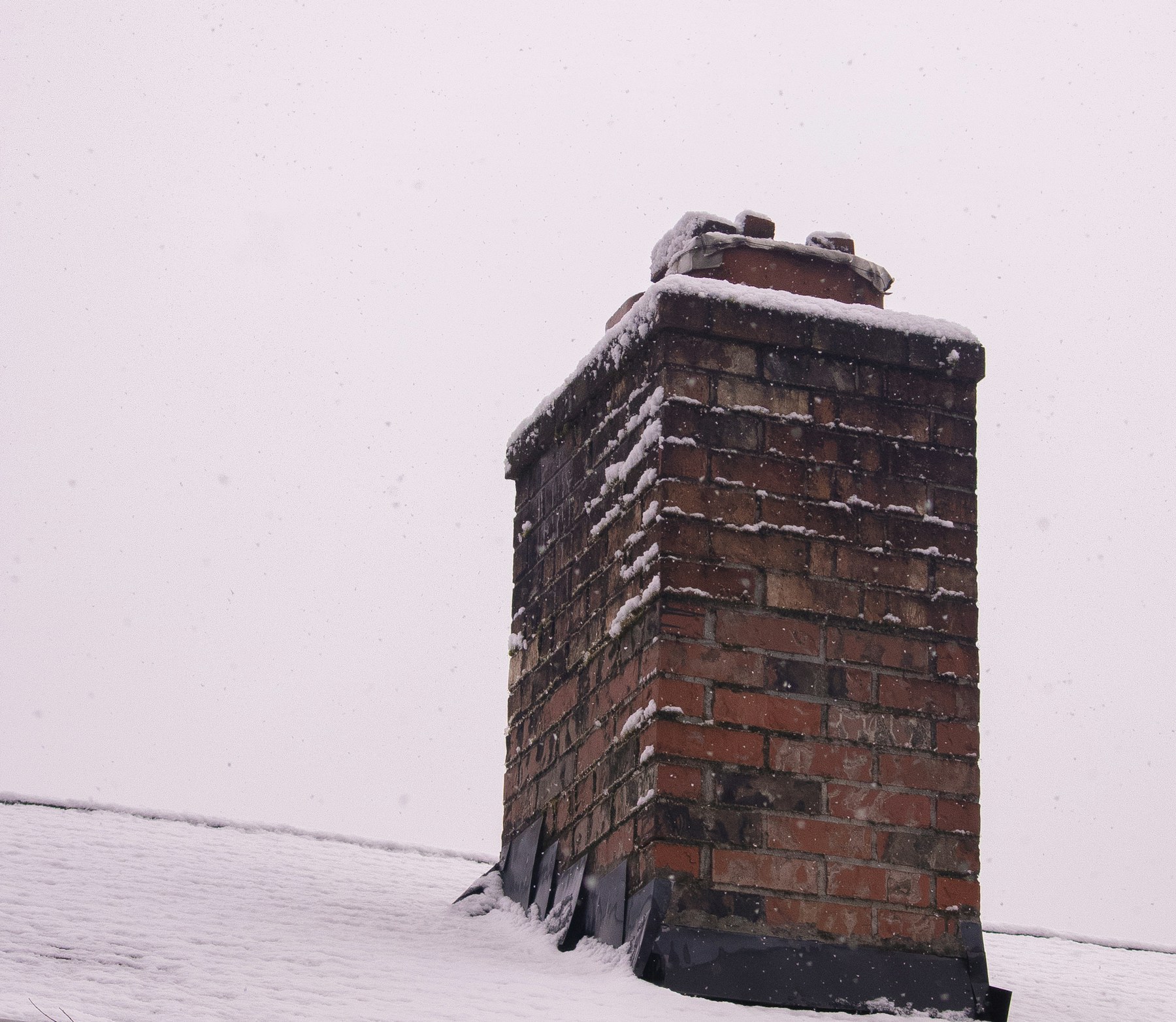 Weathered brick chimney with metal flashing on a snow covered shingle roof, the freeze and thaw exposure Coast Carpentry Home Group masons repair on Cape Cod chimneys
