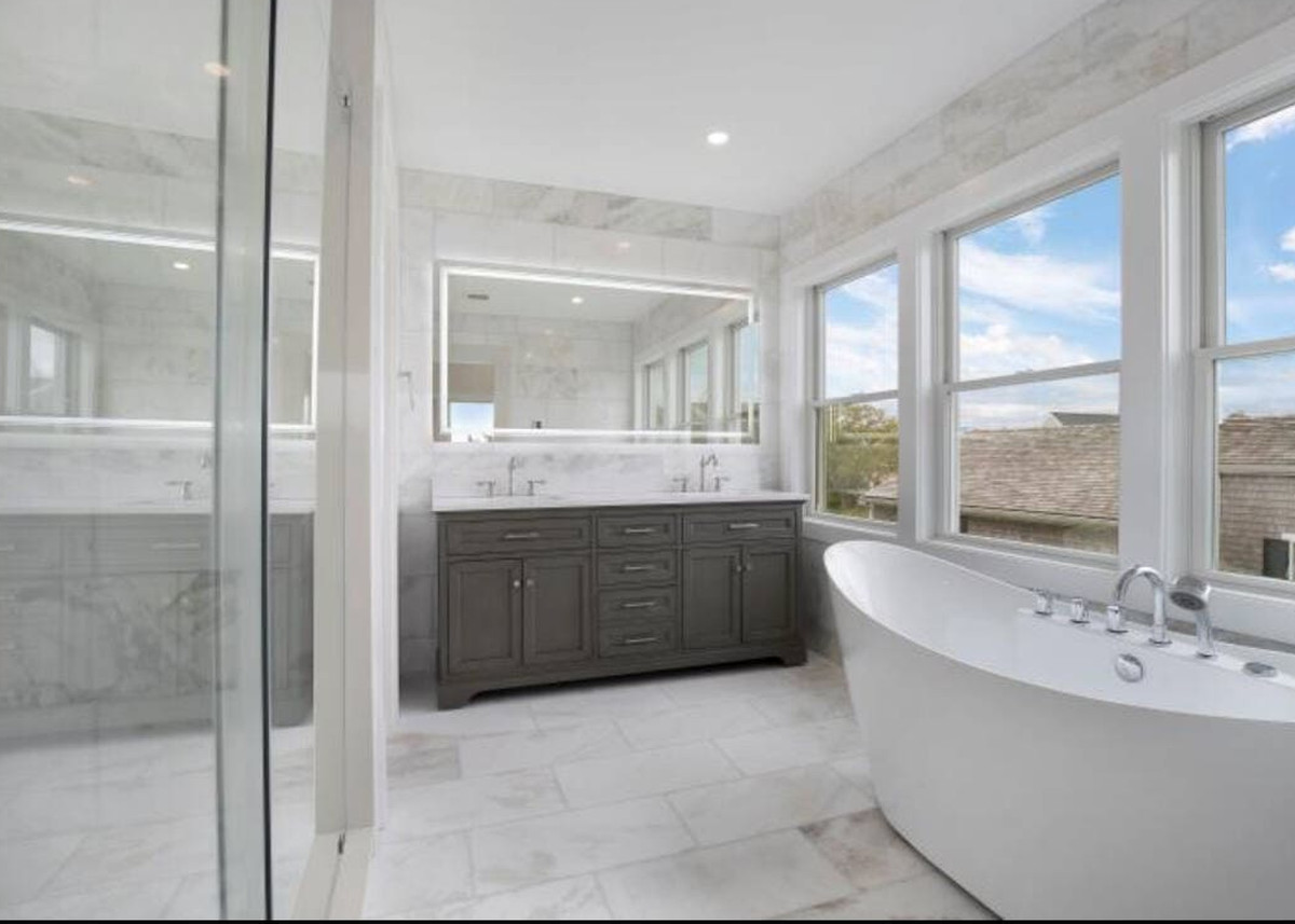 Finished primary bathroom remodel by Coast Carpentry Home Group in Hyannis, Cape Cod, with a freestanding tub under three windows, a gray double vanity, a frameless glass shower and large-format marble-look tile
