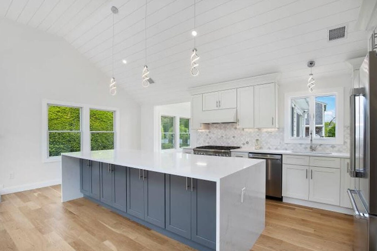 Finished kitchen by Coast Carpentry Home Group on Cape Cod with a long gray shaker island, quartz waterfall countertop, white cabinetry with crown molding and a vaulted tongue-and-groove ceiling