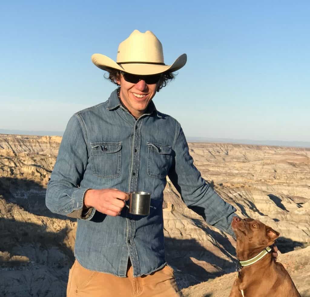 Man in cowboy hat and sunglasses with dog in desert landscape.