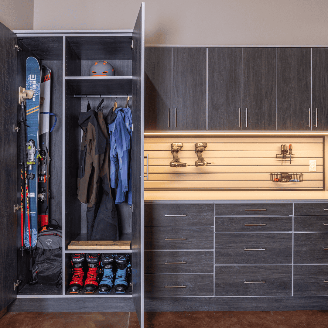 Custom dark wood garage cabinetry with an open ski locker storing skis, boots, helmets and winter jackets beside an illuminated tool-storage workspace.