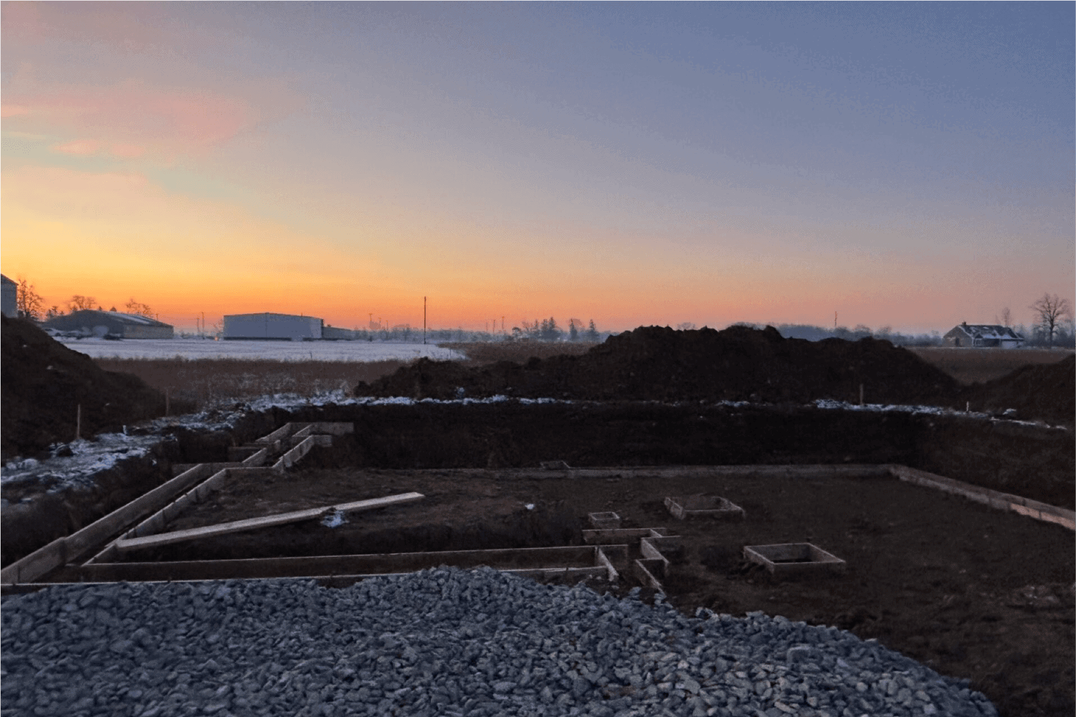 A construction site at sunrise, with early stages of foundation work in progress. Wooden formwork outlines the structure’s base, and a gravel bed is spread in the foreground. Snow lightly covers the ground, and mounds of excavated earth surround the site. In the distance, industrial buildings and a farmhouse are visible against a gradient sky of warm oranges and purples.