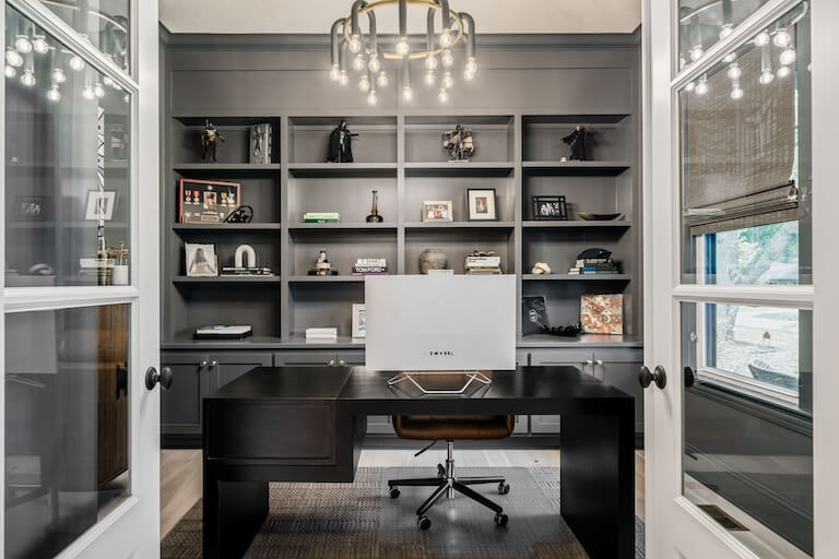 A luxury home office by Compass Homes. Open glass doors showcase a dark wood desk in the center of the room with a modern, stunning light fixture above it. Behind the desk is a wall of built-in shelving displaying an array of memorabilia, books, and picture frames.