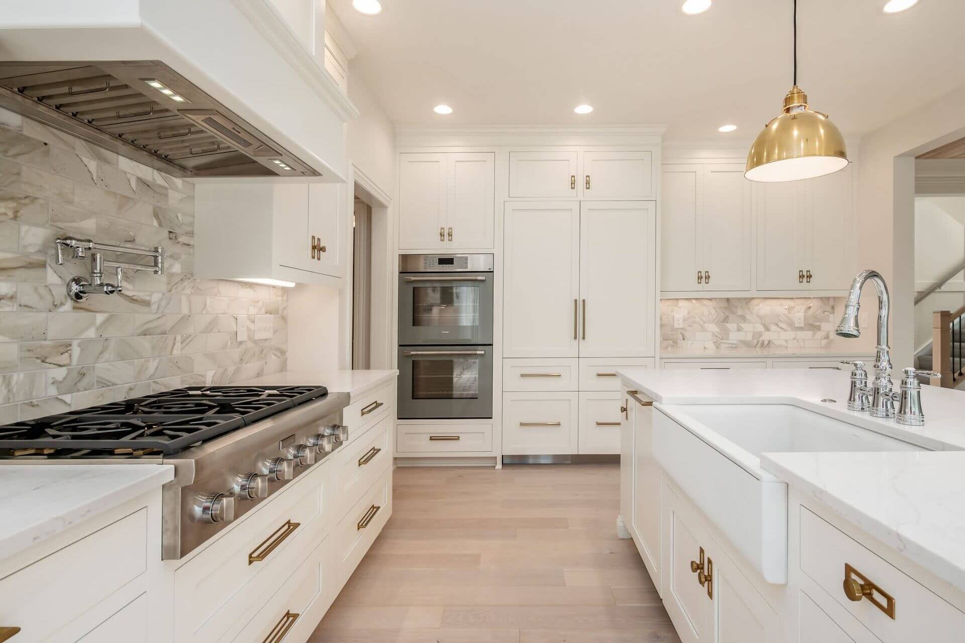 A custom, luxury Compass Homes kitchen with white cabinetry, a large white farmhouse sink with silver a faucet, a gold lighting fixture above the sink.