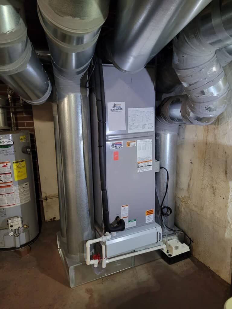 A home heating and cooling system by JP Heating & Cooling is set up in a basement with concrete floors, ready to keep the house comfortable. A water heater sits nearby.