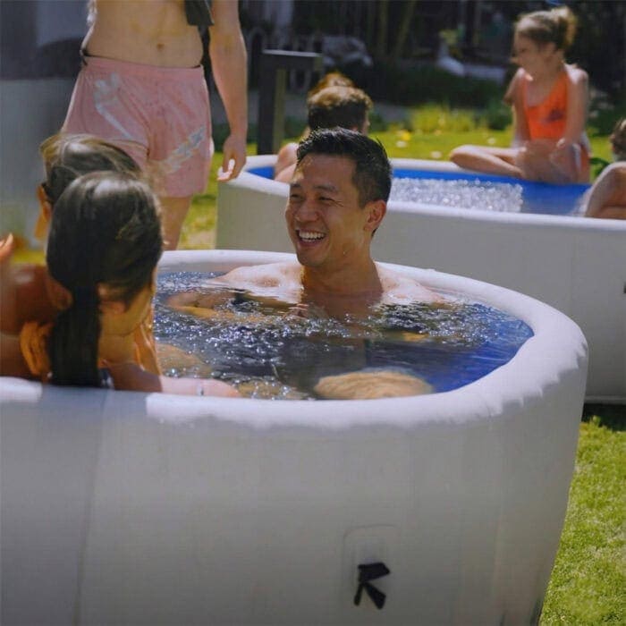 Smiling man immersed in an inflatable ice bath at a backyard group session with others in tubs nearby