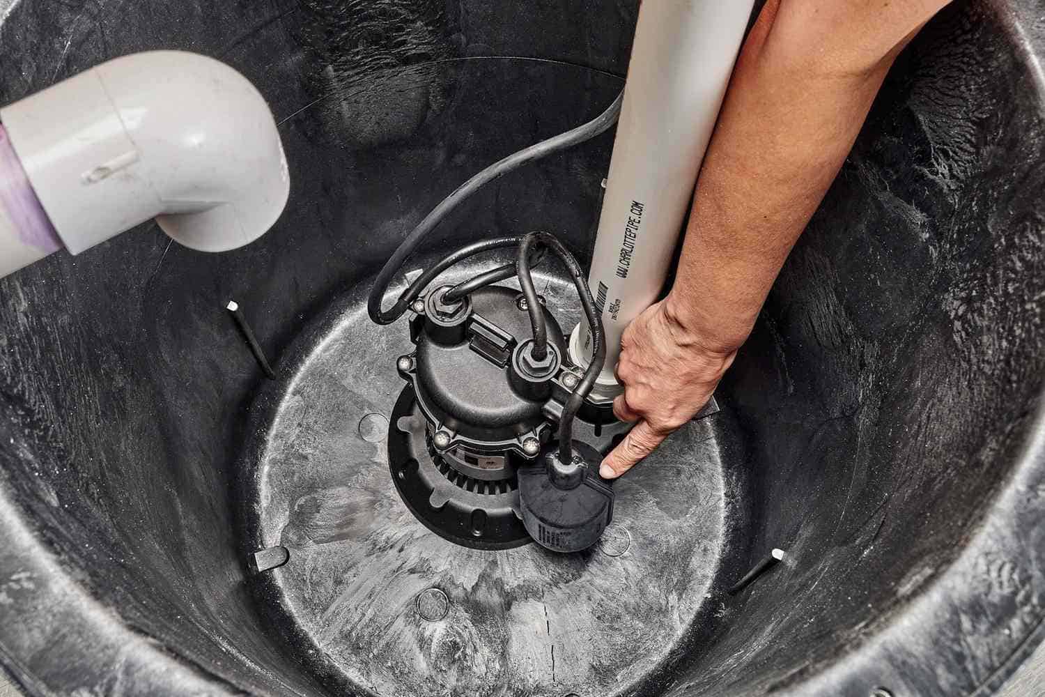 Emergency plumbing repair technician installing submersible sump pump in a black plastic basin for basement water drainage.