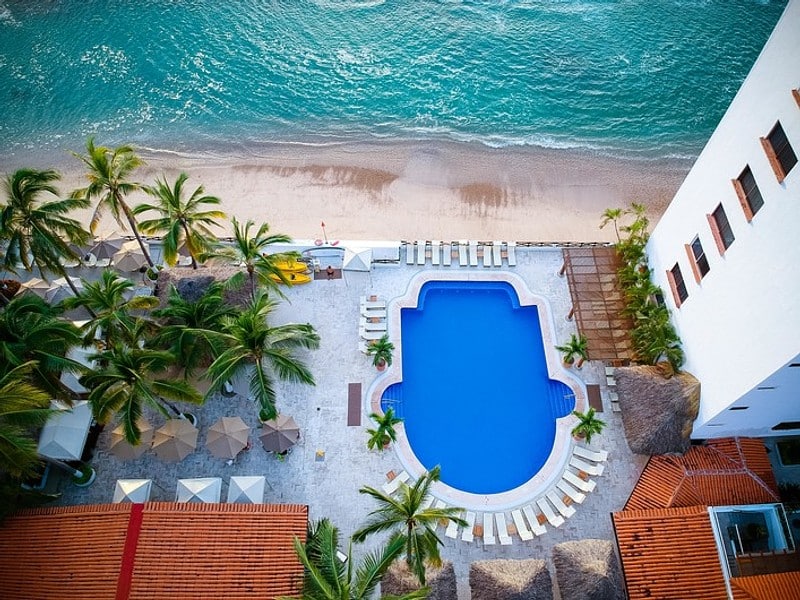 Aerial view of a hotel pool surrounded by palm trees, with a sandy beach and ocean waves in the background.