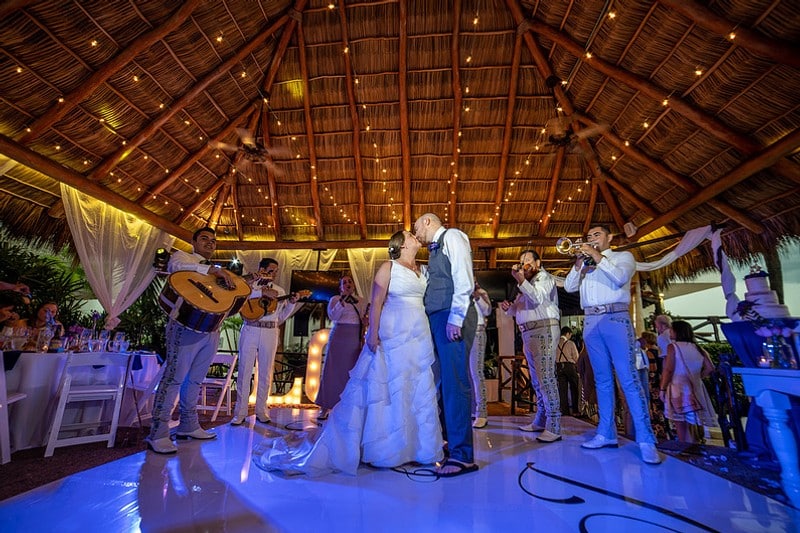 The bride and groom share a kiss on the dance floor, surrounded by soft lighting and joyful guests.