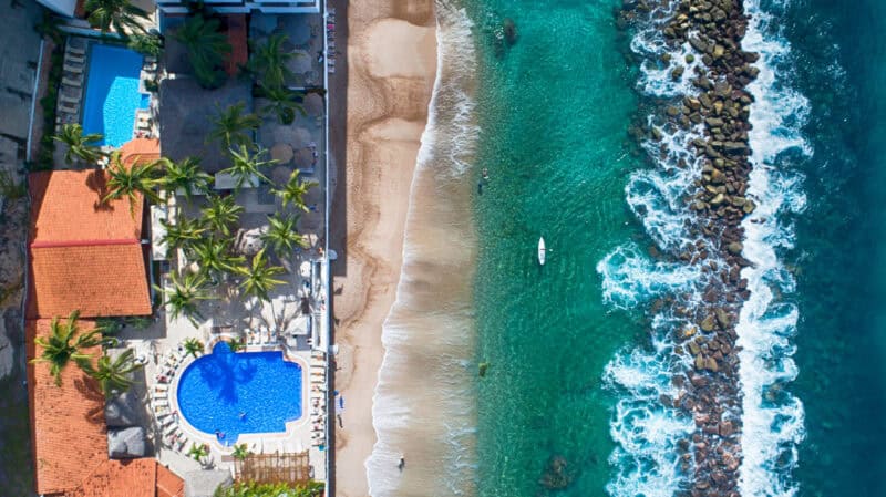 Aerial view of a beach with golden sand and a nearby hotel, showcasing the vibrant blue ocean and lush surroundings.