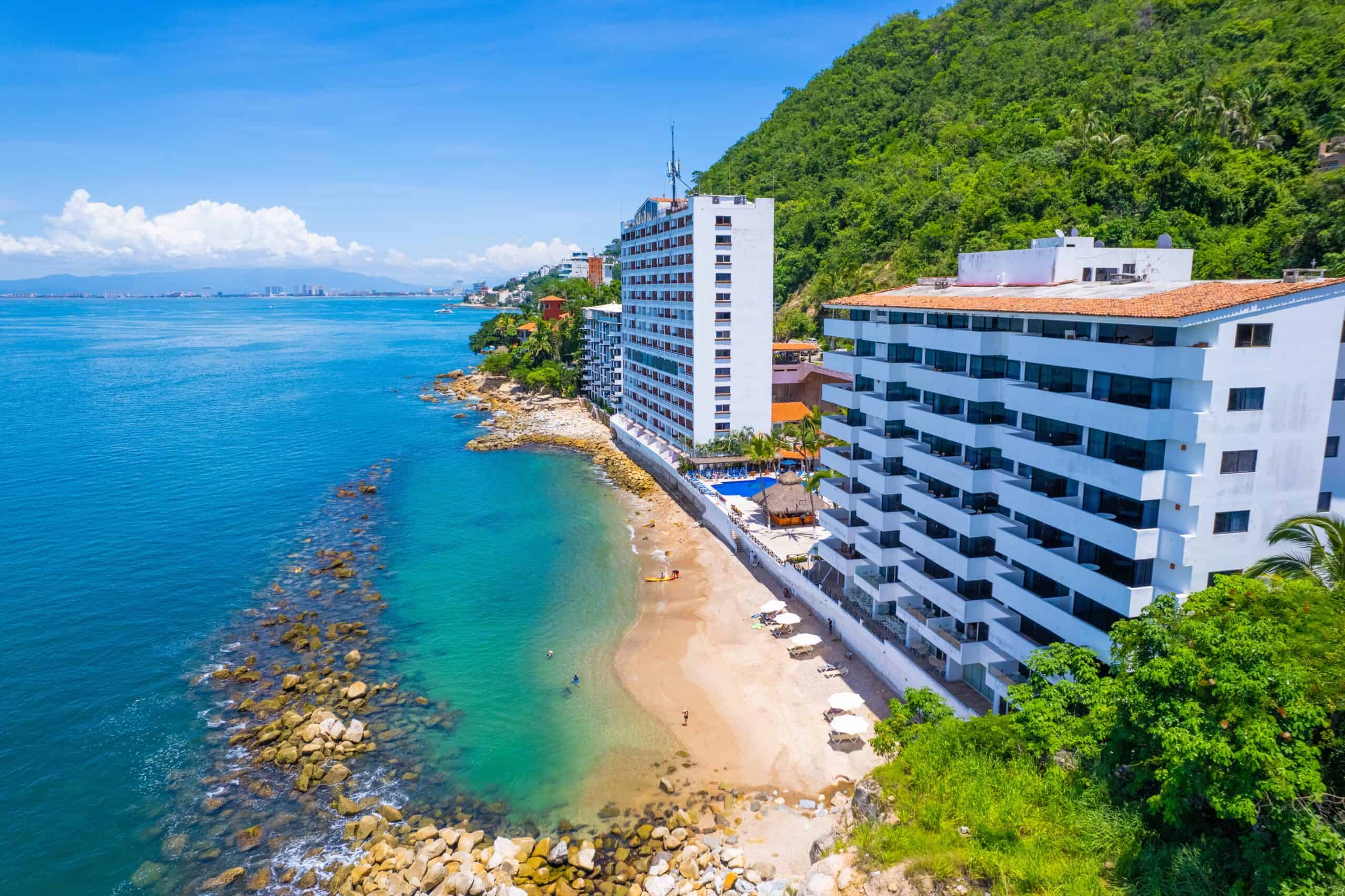 A scenic view of a beach with a hotel in Puerto Vallarta, showcasing golden sand and clear blue waters.