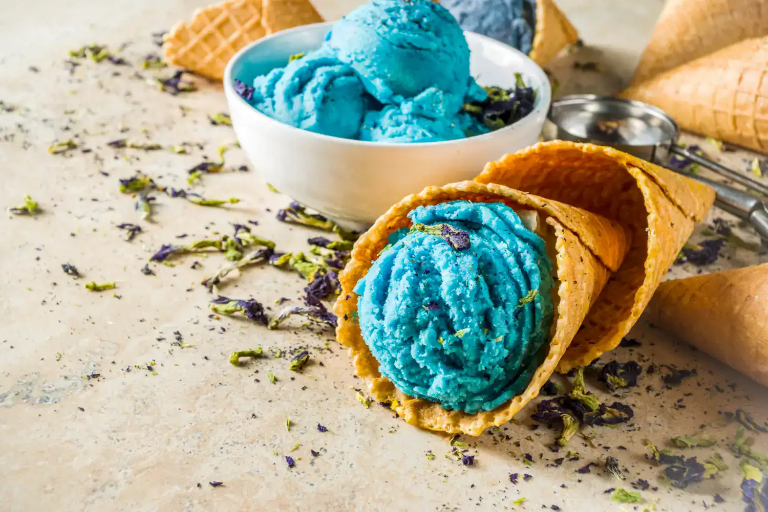 Blue Cookie Monster ice cream in cone with bowl of scoops on textured surface.