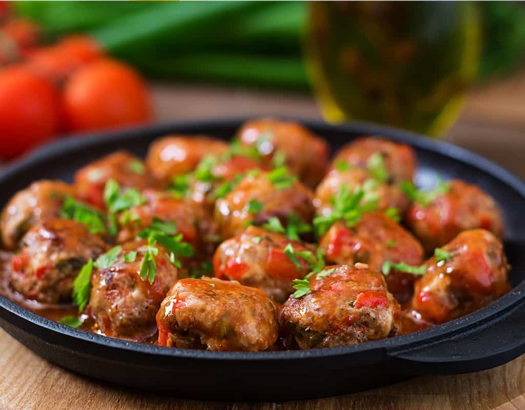 Meatballs in spicy curry sauce served on a black skillet with fresh herbs and tomatoes in the background.