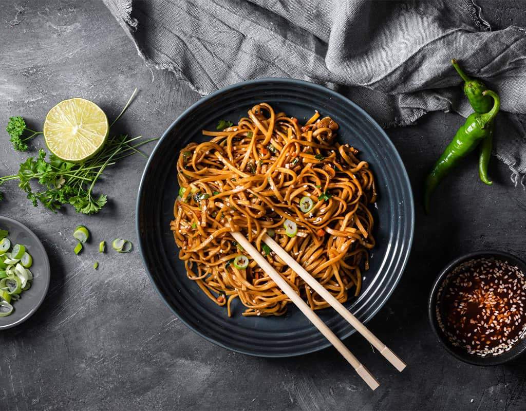 Stir-fried Indian noodles with vegetables and spicy sauce served on a dark plate.