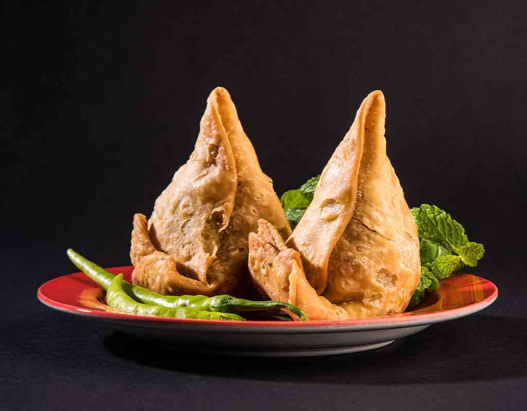 Fried Indian samosas with green chili and mint leaves on a red plate.