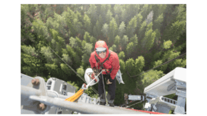 Climber in red jacket and helmet ascending a tall telecommunications tower.