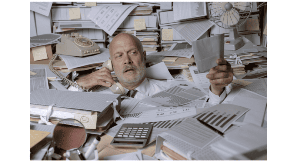 Overwhelmed man surrounded by cluttered papers and office supplies.