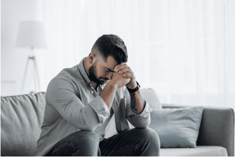 A stressed man sitting on a sofa with his head in his hands, depicting anxiety, stress, and emotional exhaustion.