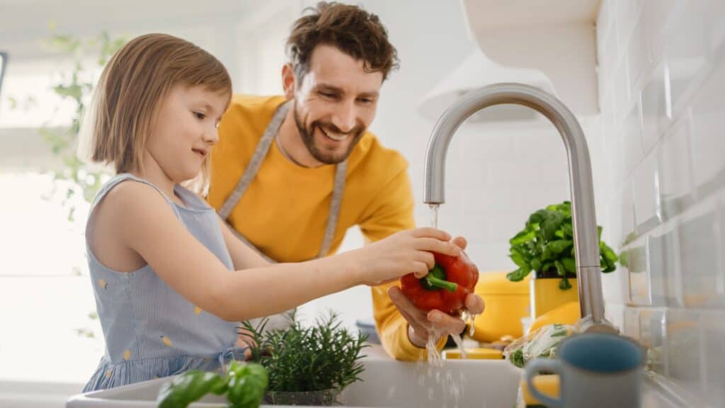 A man and a young girl wash a red bell pepper under running water in a kitchen sink surrounded by fresh herbs and vegetables, while learning tips for proper garbage disposal maintenance.