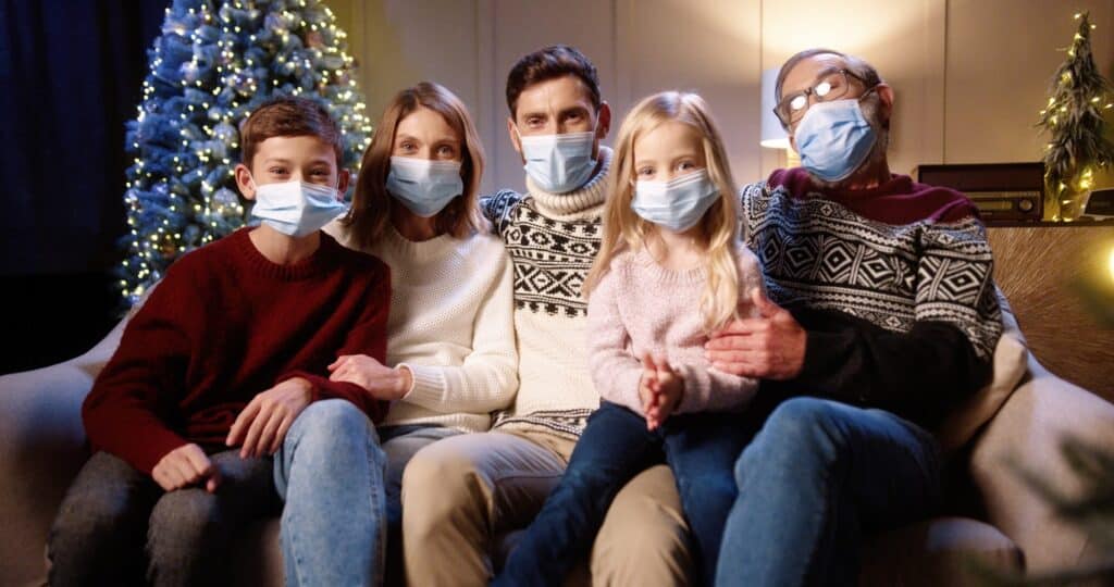Five people wearing face masks sit together on a couch in front of a decorated Christmas tree, posing for a photo and practicing holiday safety during their festive gathering.