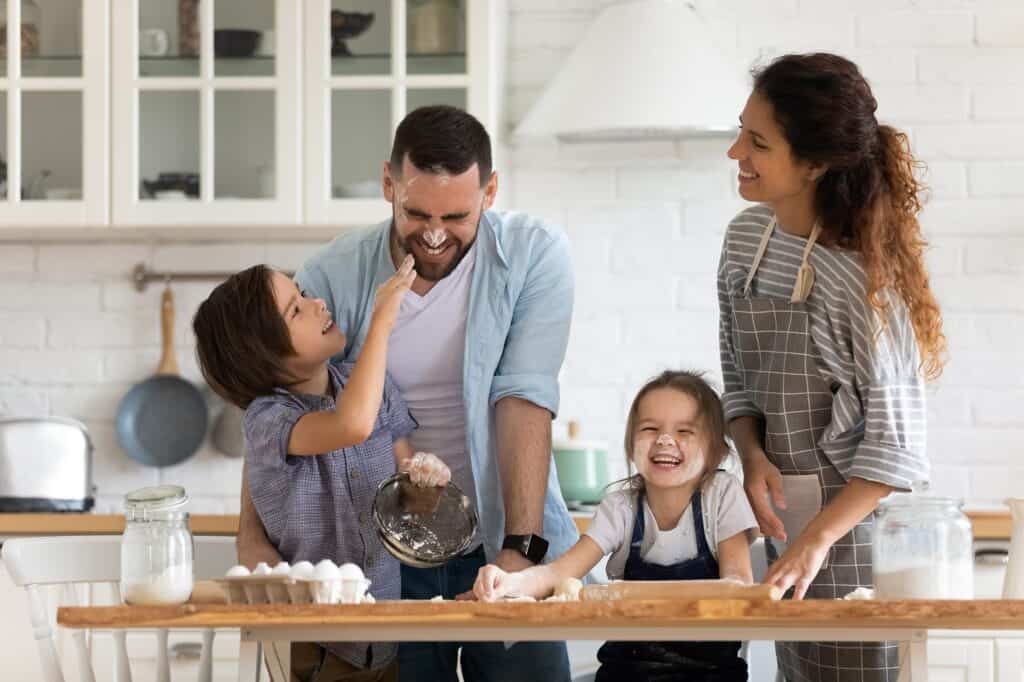 A family of four smiles and laughs together while baking in a bright kitchen, unaware that copper pipes beneath the floor might need repair or replace services due to a small pinhole leak. Flour and baking ingredients are visible on the wooden table.