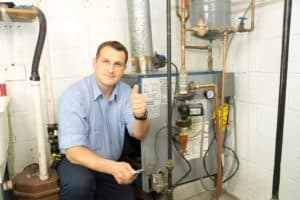 A man in a blue shirt kneels next to a boiler or furnace in a utility room, giving a thumbs up to show excellent customer service.