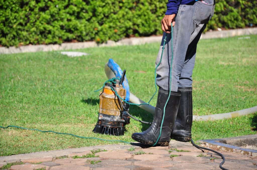 Person in rubber boots stands on grass holding a cord attached to a submersible sump pump, with a green hedge in the background—perfect for illustrating essential homeowner tips for sump pump maintenance.