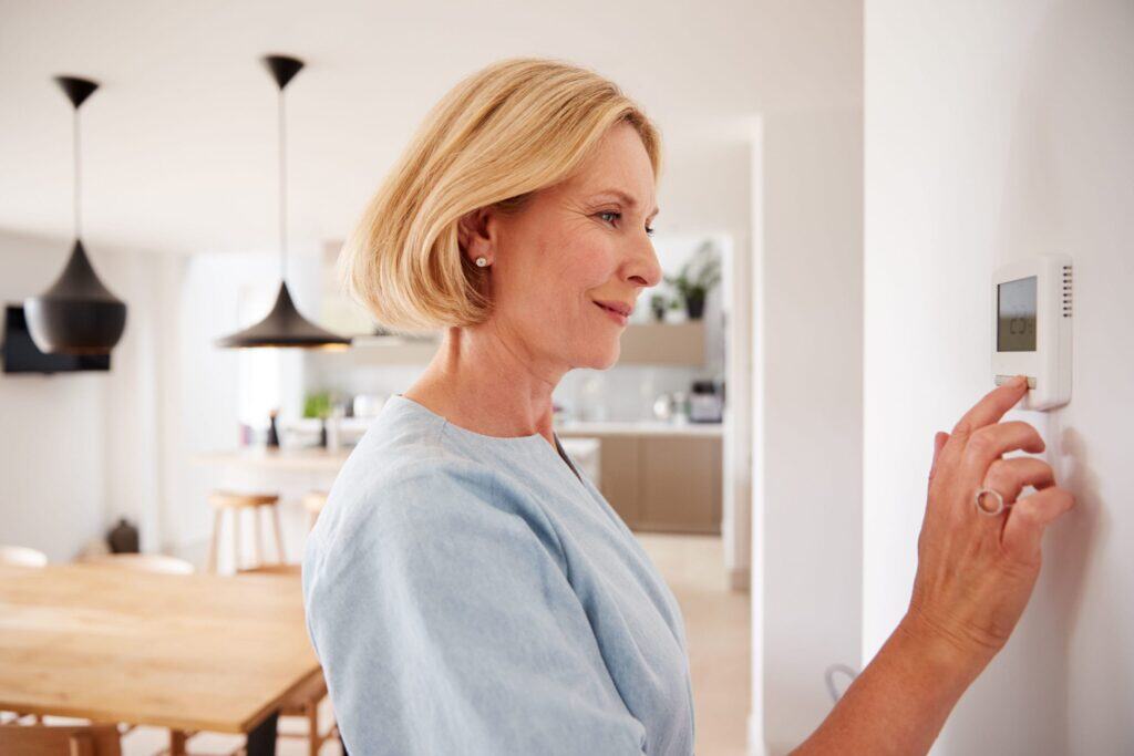 A woman adjusts a wall-mounted thermostat in a modern kitchen and dining area, checking for heating system signs that may indicate the need for heating repair.