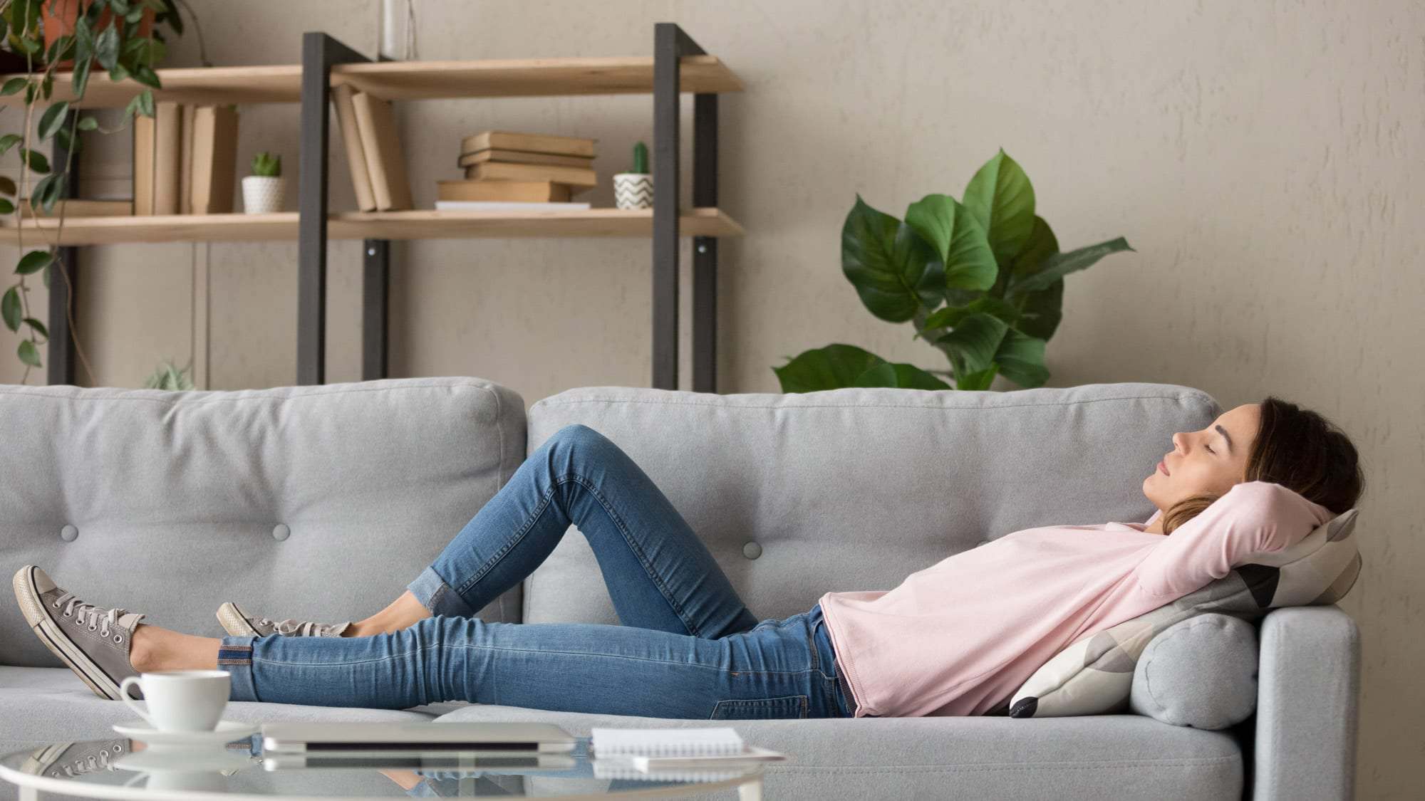 A woman in casual clothes lies on a gray couch with her eyes closed, hands behind her head, in a living room with shelves, plants that help improve air quality, and books in the background.