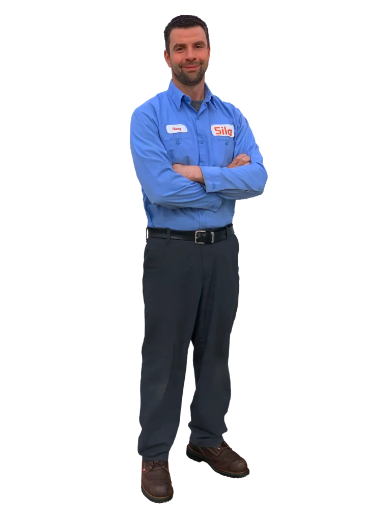 A man wearing a blue work shirt with a "Sila" logo and name tag, dark pants, and brown shoes stands with arms crossed, smiling.
