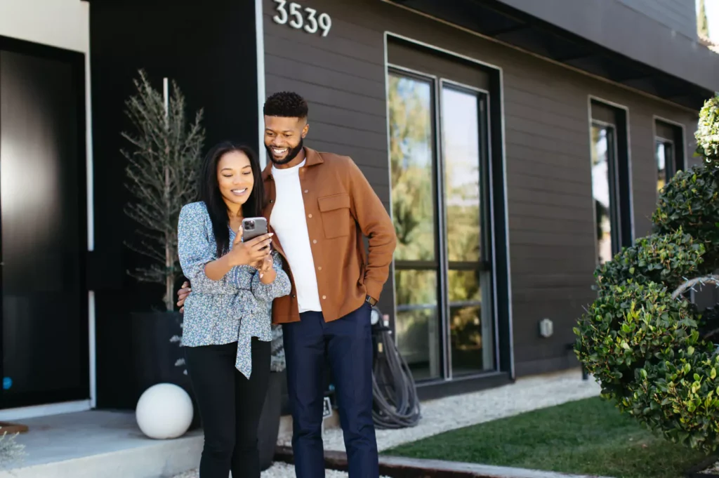 A couple stands outside a modern house, smiling and looking at a smartphone together.