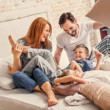A family of four sits on a bed, laughing and playing with pillows in a bright, cozy bedroom cooled by high velocity air conditioning.
