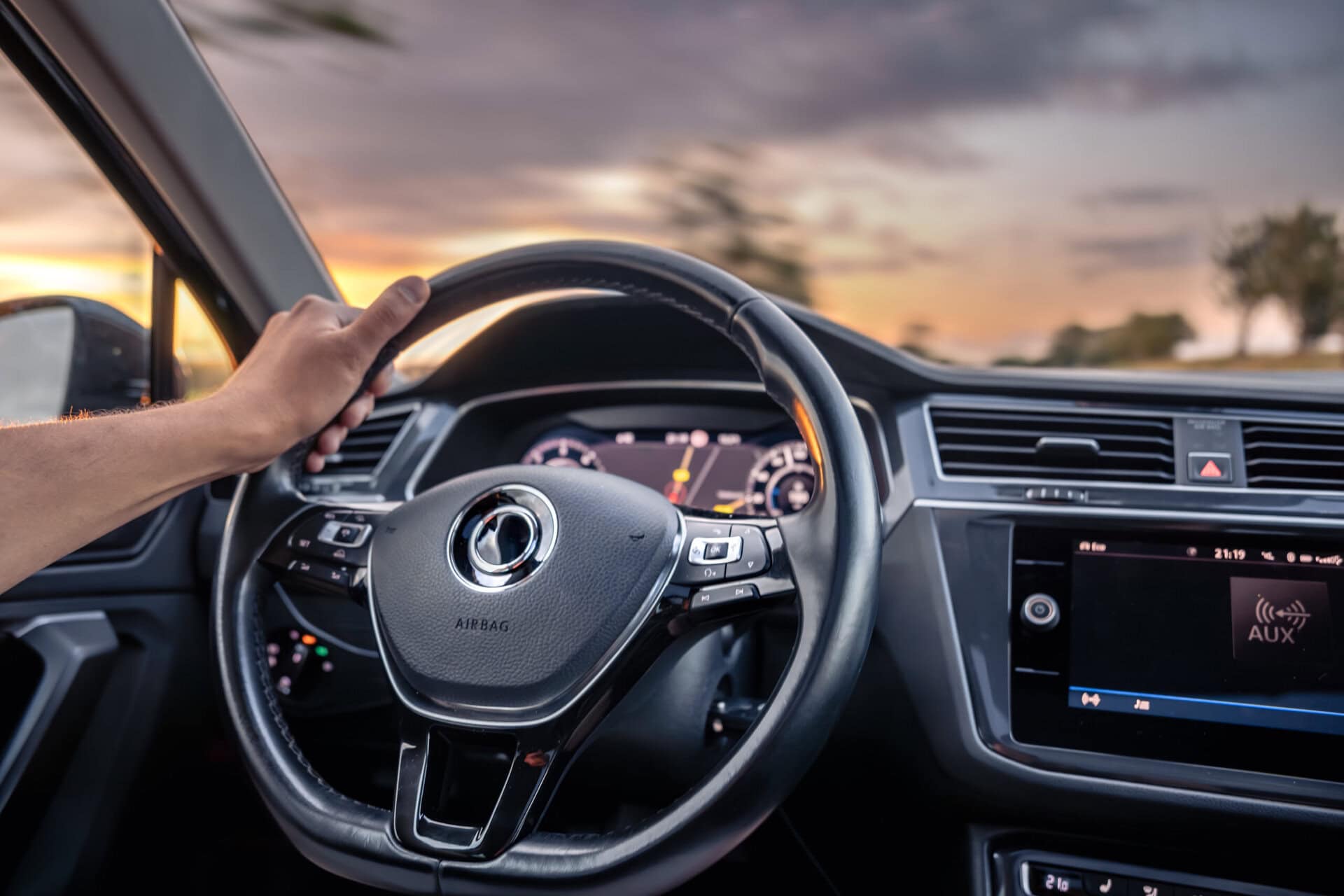Close-up of a person holding a steering wheel inside a car, highlighting suspension and steering system components during a sunset drive.