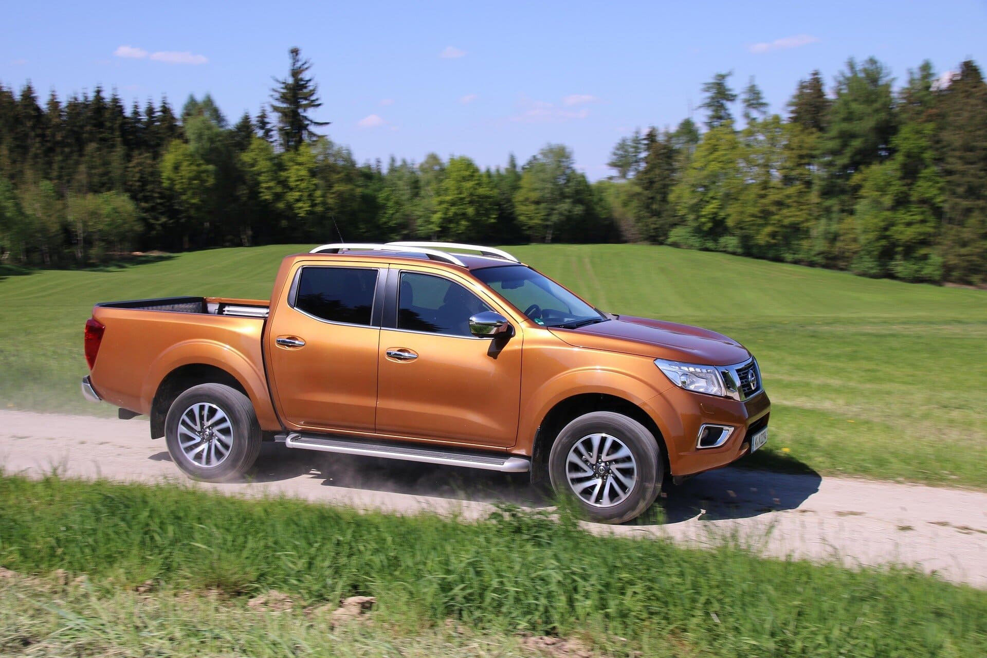 An orange Nissan pickup truck driving on a dirt path surrounded by lush green grass and trees, showcasing durability and off-road capability.