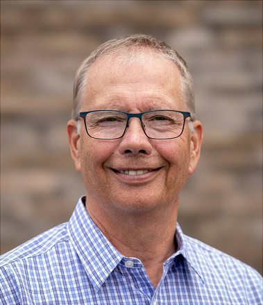 Tom Deverell has short light brown hair and glasses, smiling at the camera, wearing a blue and white checked shirt.