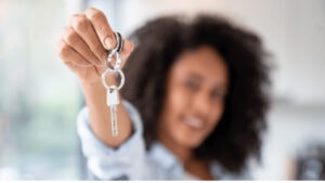 Key holder with a silver key in focus, smiling woman with curly hair blurred in the background, real estate agent promoting property sales, Dallas Fort Worth real estate expert.