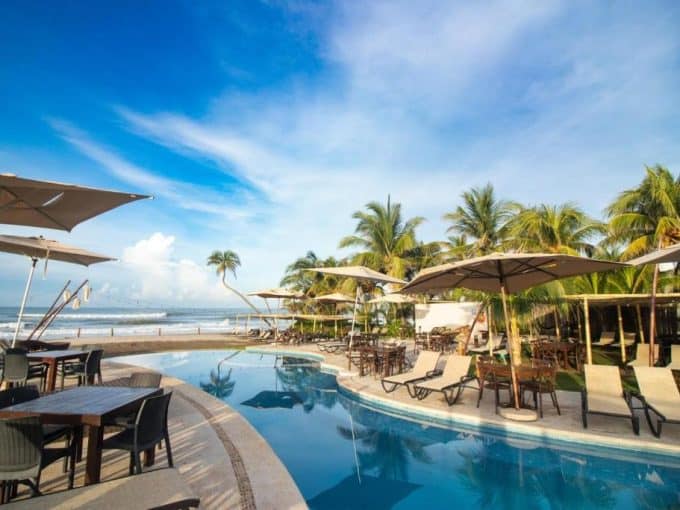 Luxurious Swimming Pool At Diamante Cabo San Lucas Beachfront Resort With Palm Trees, Sun Loungers, And Seaside Views, Ideal For Relaxation And Upscale Vacation Stays.