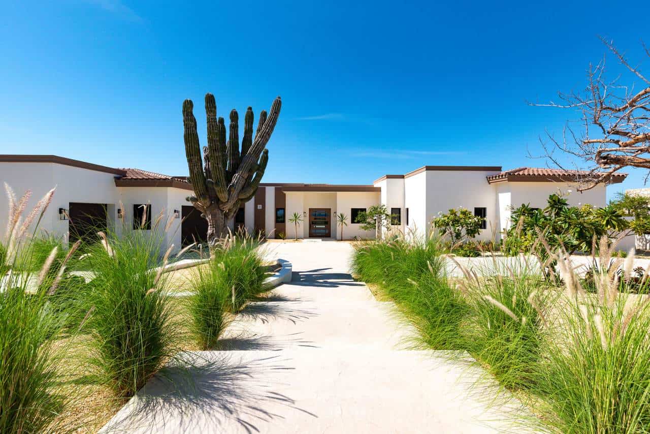 Luxurious Modern Home In Diamante Cabo San Lucas Featuring Desert Landscaping With Cacti And Lush Green Plants, Under A Clear Blue Sky. Diamante Cabo San Lucas Offers Premium Real Estate Options.