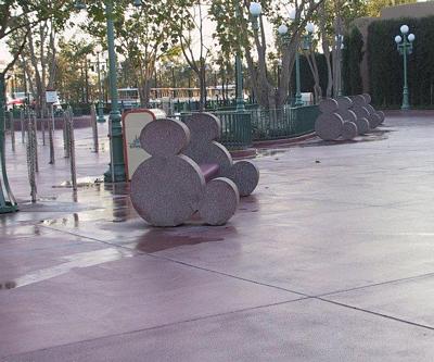 Whimsical Mickey-shaped benches line the path in the Gate Area, nestled among trees and classic Disney park lampposts.