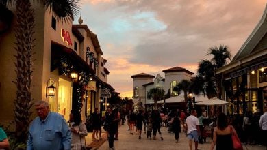 Bustling promenade at sunset with visitors strolling past palm trees, lively shops, and vibrant entertainment venues.