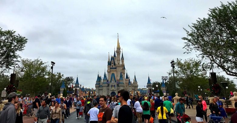 A large crowd of people walk toward Cinderella Castle at a Disney park on a cloudy day, with trees and decorative lampposts lining the pathway.