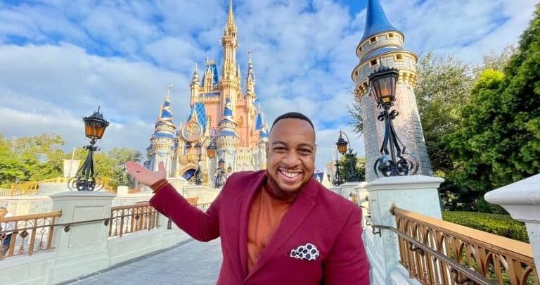 Guest in a maroon suit smiles for a photo in front of the iconic fairytale castle at a bustling amusement park.