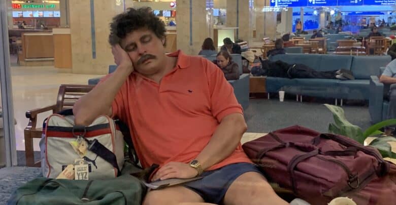 A traveler in a red polo shirt naps near his luggage in the airport, catching some rest before flying to the theme park merger event.