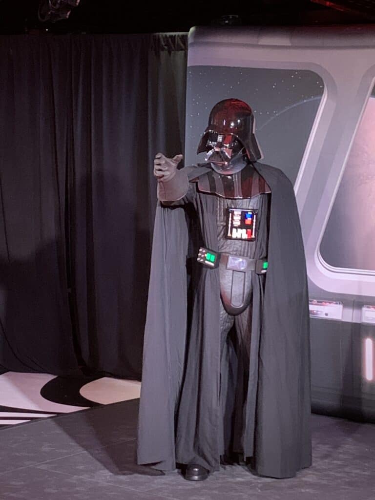 Darth Vader aboard The Disney Fantasy poses indoors, extending a gloved hand toward guests for an epic Star Wars encounter.