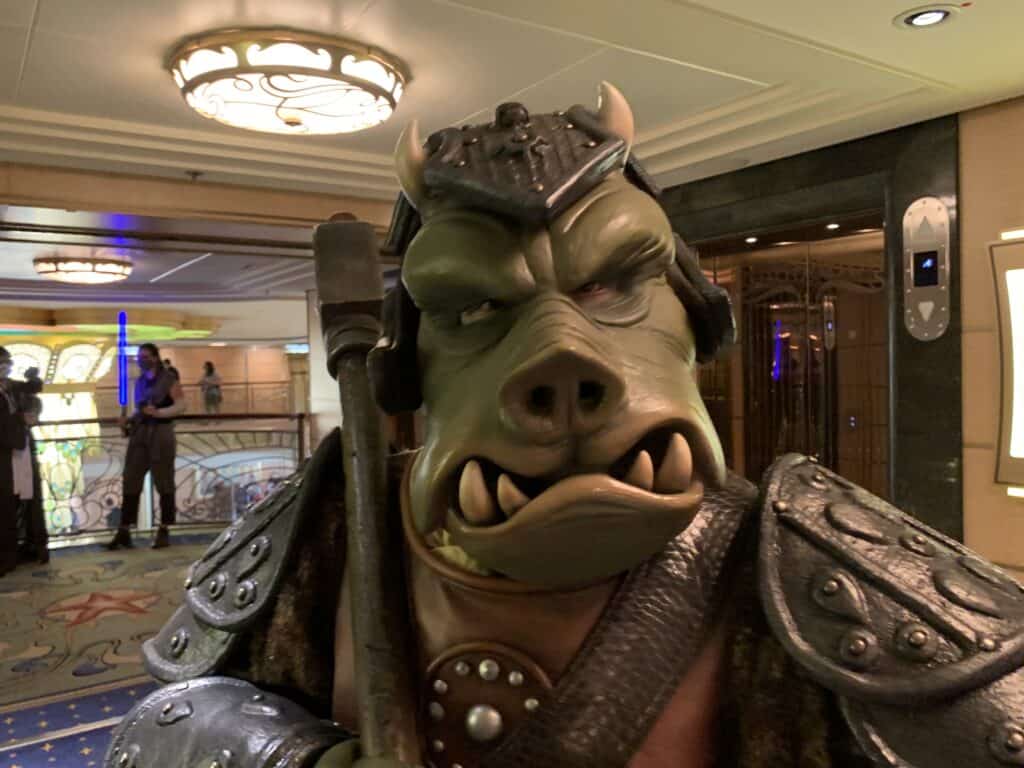 Gamorrean Guard greets guests with a staff in a themed hallway aboard Disney Fantasy, ready for photos and fun.