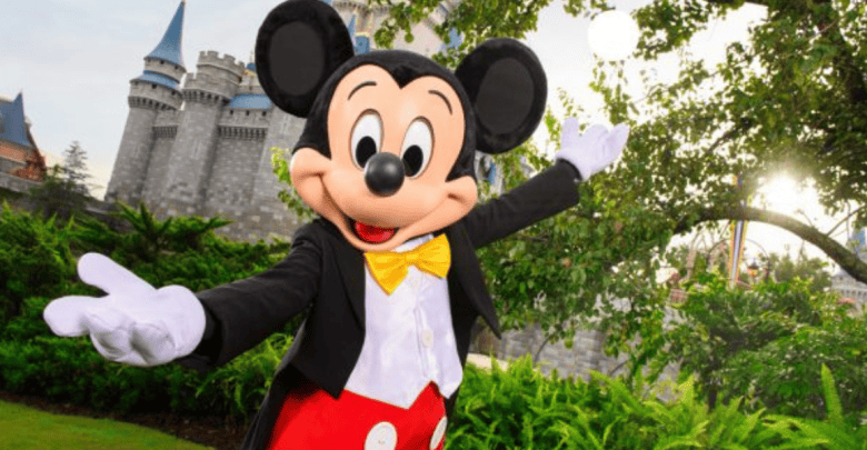 A person in a Mickey Mouse costume stands joyfully with open arms in front of a castle surrounded by greenery. Mickey is wearing a black tuxedo jacket, yellow bow tie, and red shorts with white buttons. The sun shines brightly in the background.