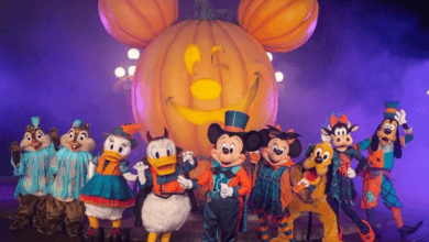 A group of Disney characters, including Mickey Mouse, Donald Duck, Goofy, and Pluto, dressed in Halloween costumes, stand in front of a large illuminated pumpkin with Mickey Mouse's face carved into it. The background is filled with purple mist.